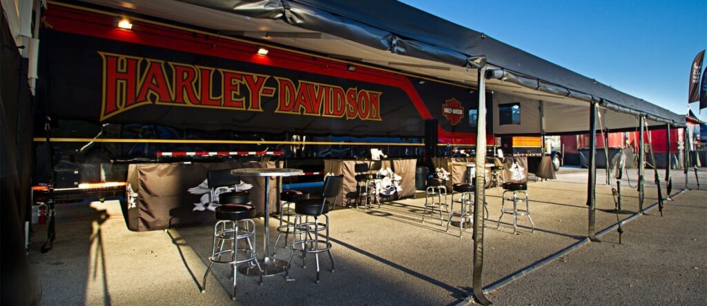 A Harley-Davidson sign is prominently displayed above a bar area with stools and tables under a clear blue sky.