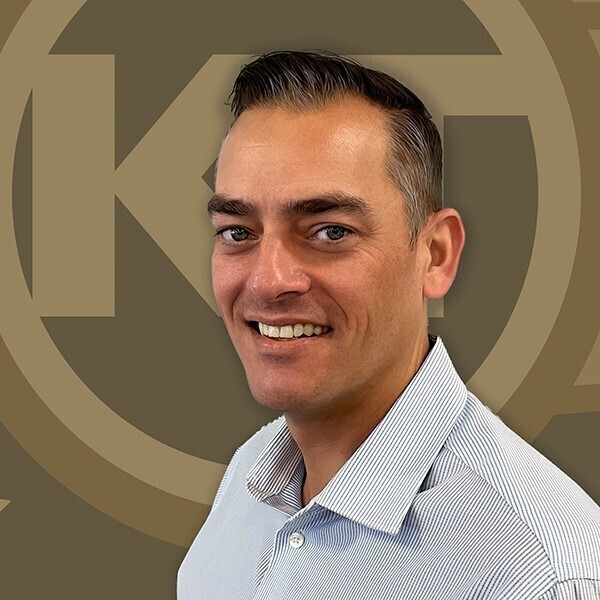 A smiling individual with a collared shirt stands against a background featuring a circular logo with letters inside it.