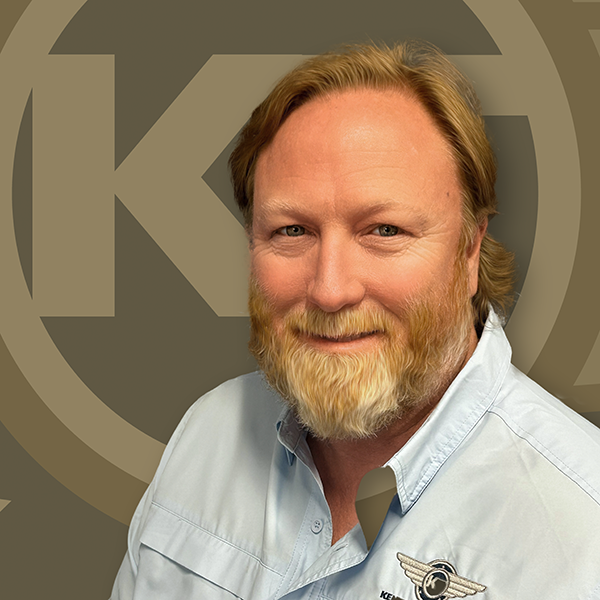 A smiling individual with a beard is wearing a light blue shirt in front of an emblem with interlocking circles.