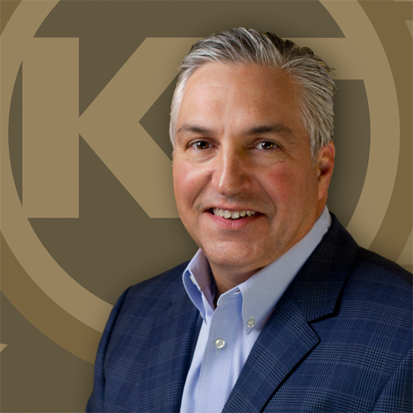 A smiling individual is posing in front of a background with a monogram logo, dressed in a professional blue shirt and blazer.