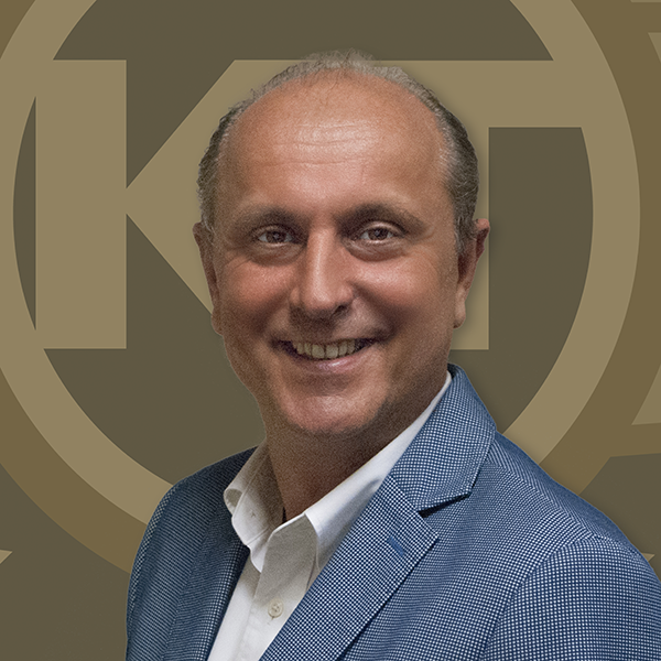 A smiling individual wearing a blue jacket stands in front of a background featuring a large, circular gold-colored logo with an intricate design.