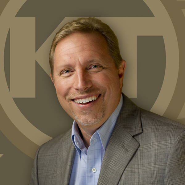 A smiling individual wearing a suit poses in front of a background with a circular logo.