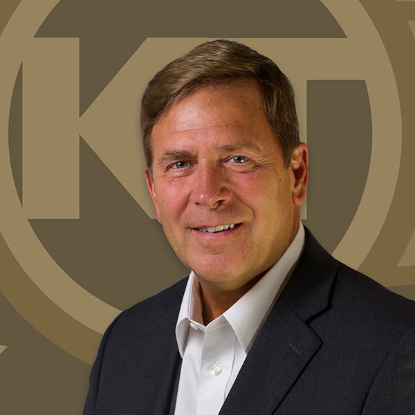A smiling individual is posed against a background featuring a circular emblem with intertwined letters.
