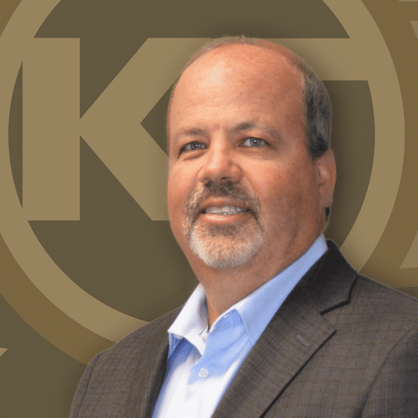 A smiling individual with a mustache is wearing a suit and blue shirt against a backdrop featuring a circular emblem with intertwined letters.