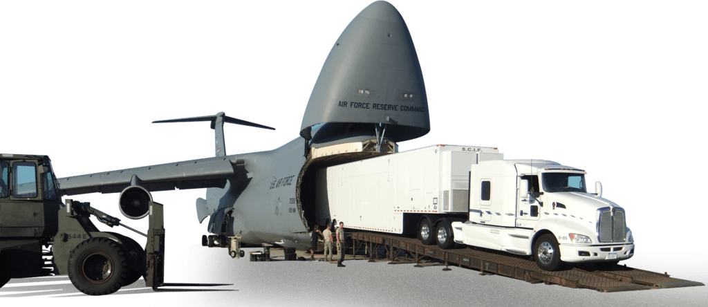 A large military cargo aircraft is being loaded or unloaded with a white semi-trailer truck backed up to its open cargo bay.